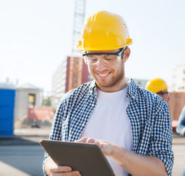 White male with yellow hard hat and safety glasses, on a construction site smiling while looking at clipboard
