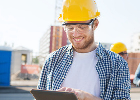 White male with yellow hard hat and safety glasses, on a construction site smiling while looking at clipboard