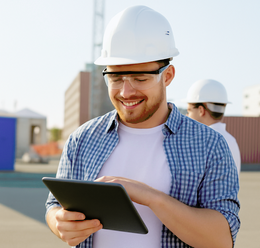 White male with yellow hard hat and safety glasses, on a construction site smiling while looking at clipboard