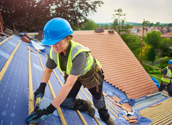 white roofer wearing black gloves, hi vis and blue hard hot installing wooden battens on a roof
