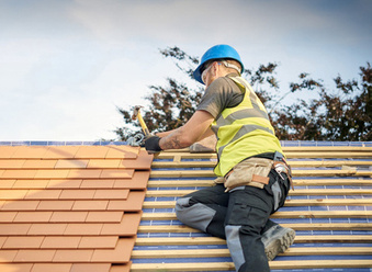 A roofing contractor in hi vis and blue hard hat laying small roof tiles on a pitched roof