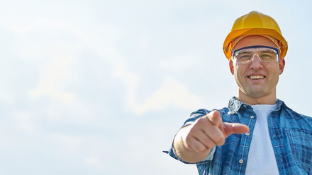 white male contractor wearing white t shirt, blue checked shirt, yellow hard hat and safety glasses pointing forwards